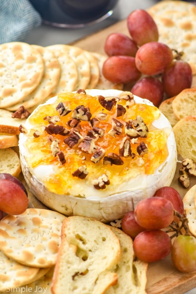 wheel of brie in tact with honey, marmalade, pecans on a platter with crackers, baguettes, and grapes