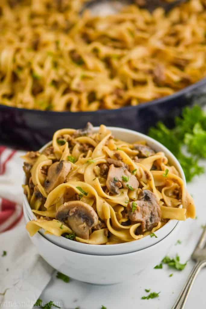 two stacked white bowls with the top one full of ground beef stroganoff recipe - egg noodles, mushrooms, parsley, and a skillet with the rest of the recipe in the background