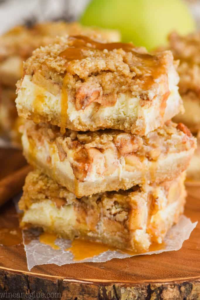a stack of three apple cheesecake bars on wax paper on a wood platter with caramel sauce dripping from them and a bite out of the top one