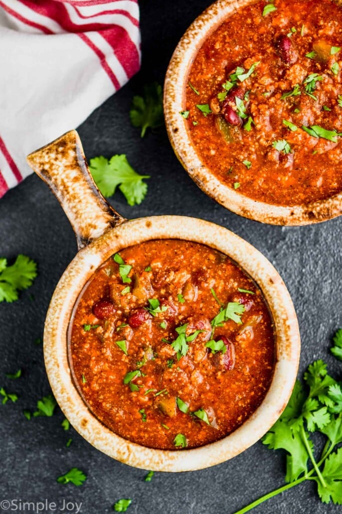 overhead view of a brown bowl with a handle holding vegetarian bean chili recipe garnished with cilantro on a dark surface with more cilantro around it