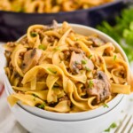 two stacked white bowls with the top one full of ground beef stroganoff recipe - egg noodles, mushrooms, parsley, and a skillet with the rest of the recipe in the background