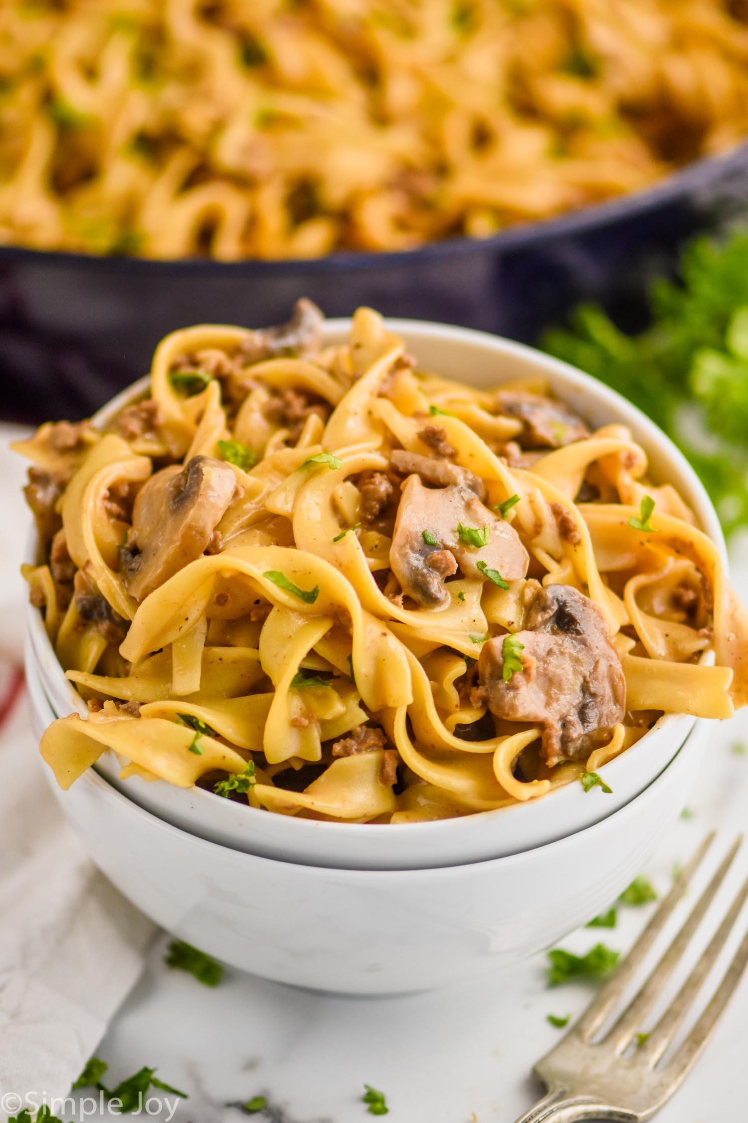 two stacked white bowls with the top one full of ground beef stroganoff recipe - egg noodles, mushrooms, parsley, and a skillet with the rest of the recipe in the background