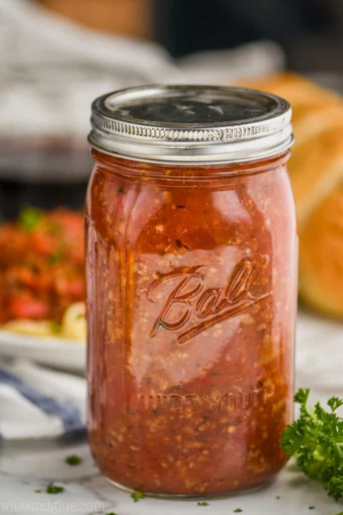 ball jar filled with meat sauce for pasta, with a plate of pasta blurred in the background and a french baguette, parsley in the foreground