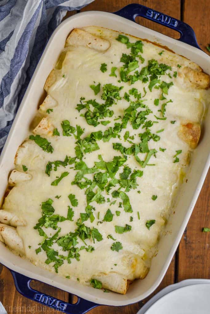 over head view of a baking dish with blue handles on a wood board holding sour cream chicken enchiladas, with a white sauce, garnished with a lot of cilantro