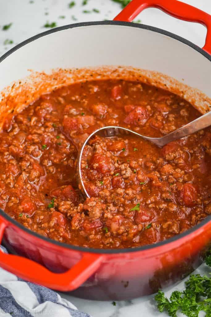 red dutch oven with spaghetti meat sauce in it, with a ladle, garnished with a little parsley