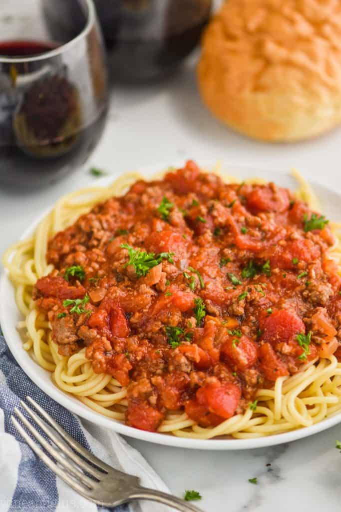 white plate with spaghetti topped with spaghetti meat sauce recipe, two red wines and bread in the background, fork in the foreground