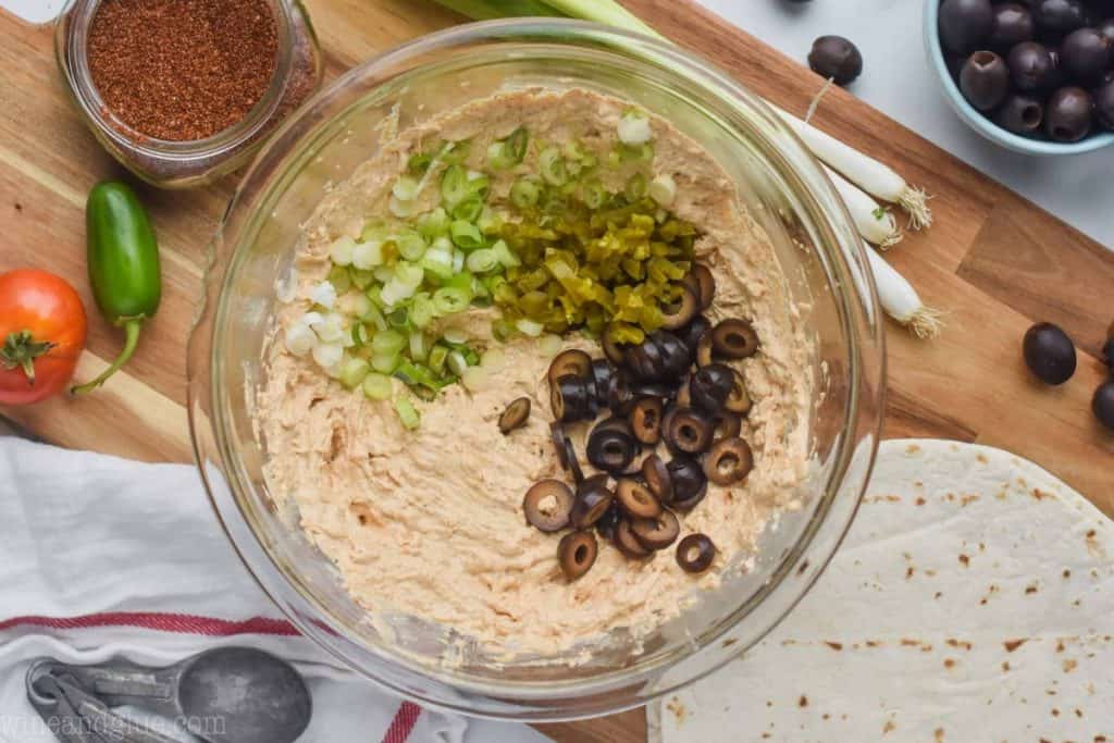 overhead view of a clear bowl with the filling for mexican pinwheels plus scallions, diced jalapeños, and sliced olives 