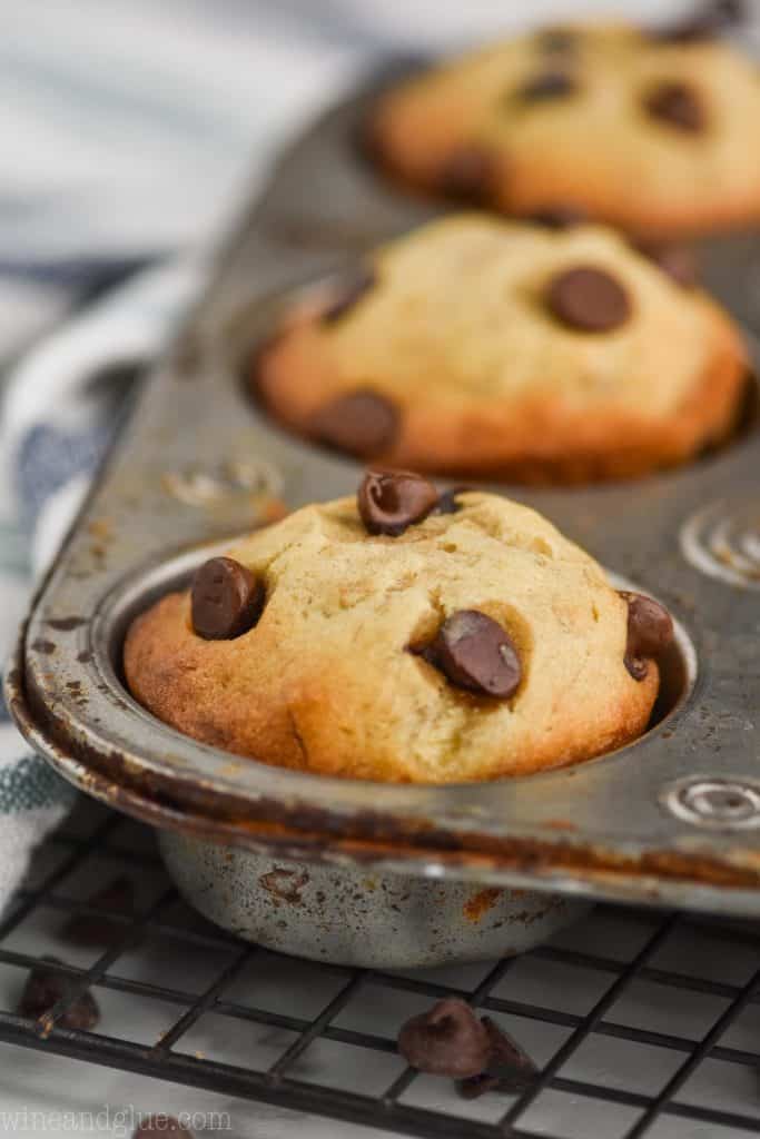 a close up of a banana chocolate chip muffin that is still in the muffin tin sitting on top of a wire cooling rack