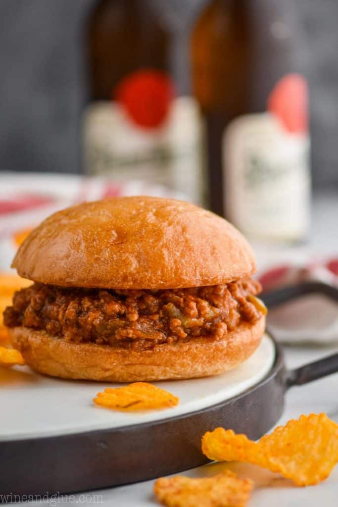 a homemade sloppy joe sitting on a metal and wood tray with chips around it and beer in the background