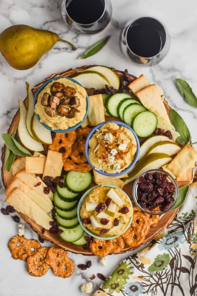 Overhead photo of a cheese board filled with sliced pears, sliced cucumbers, sliced cheese, pretzels, dried cranberries, a bowl of crispy leeks and blue cheese humus, another bowl with sage butter mushroom and humus, and a bowl of honey cranberry pear and humus. 