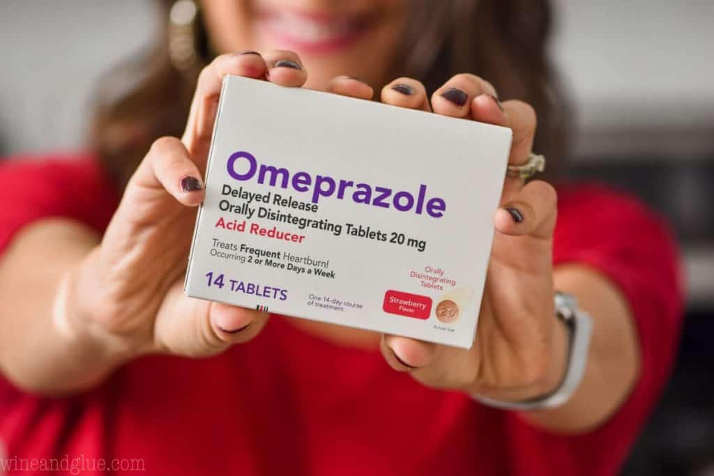 a woman's hands (with the woman blurred in the background) holding a box of omeprazole