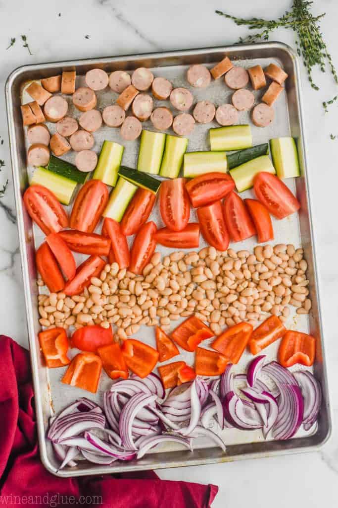 overhead view of chicken sausages, zucchini, tomatoes, northern beans, red peppers, and red onions all chopped on a rimmed baking sheet to make sheet pan sausage and veggies