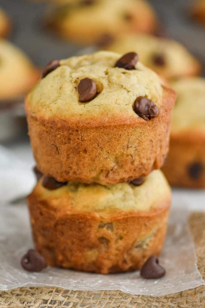 two banana chocolate chip muffins stacked on top of each other on top of wax paper with more muffins in the background