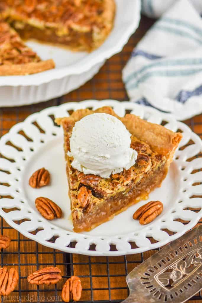 a piece of the best pecan pie on a fancy white plate with a large scoop of vanilla ice cream on top and the rest of the pie blurred in the background