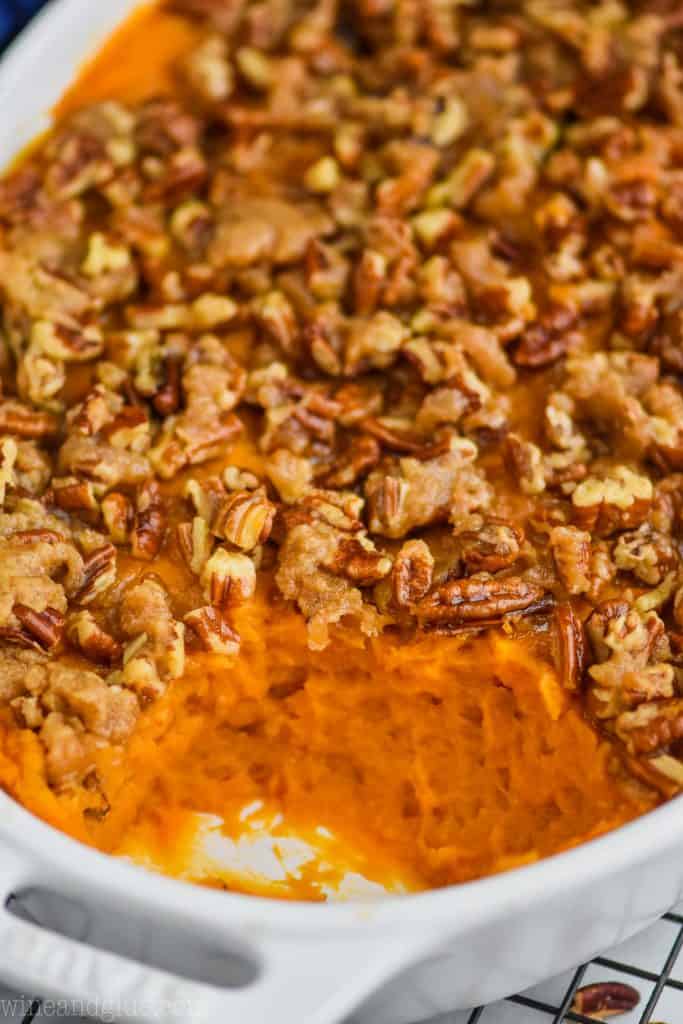 close close up of a sweet potato casserole that has been topped with pecans, butter, brown sugar, and flour and is in a white casserole dish where a scoop is missing