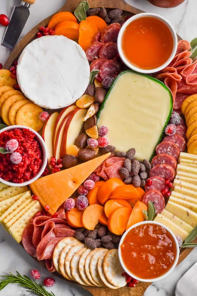 overhead view of a charcuterie board with four types of cheese, three types of crackers, persimmons, pears, oranges, and three types of spread