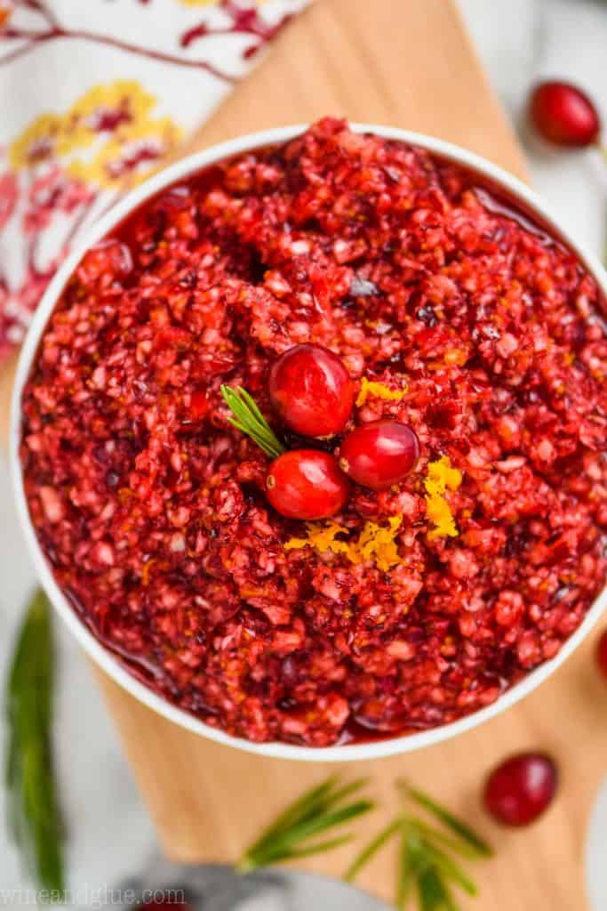 overhead view of cranberry orange relish that is in a white bowl and garnished with three cranberries, a little rosemary, and some orange zest