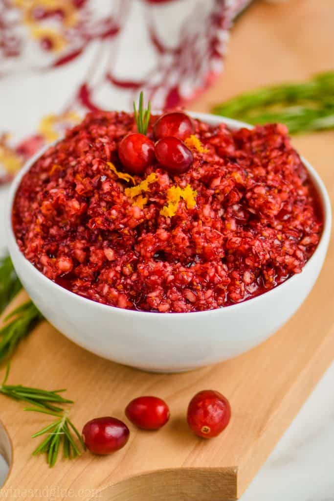 a white bowl full of cranberry relish recipe on a cutting board, garnished with three cranberries, orange zest, and a little rosemary