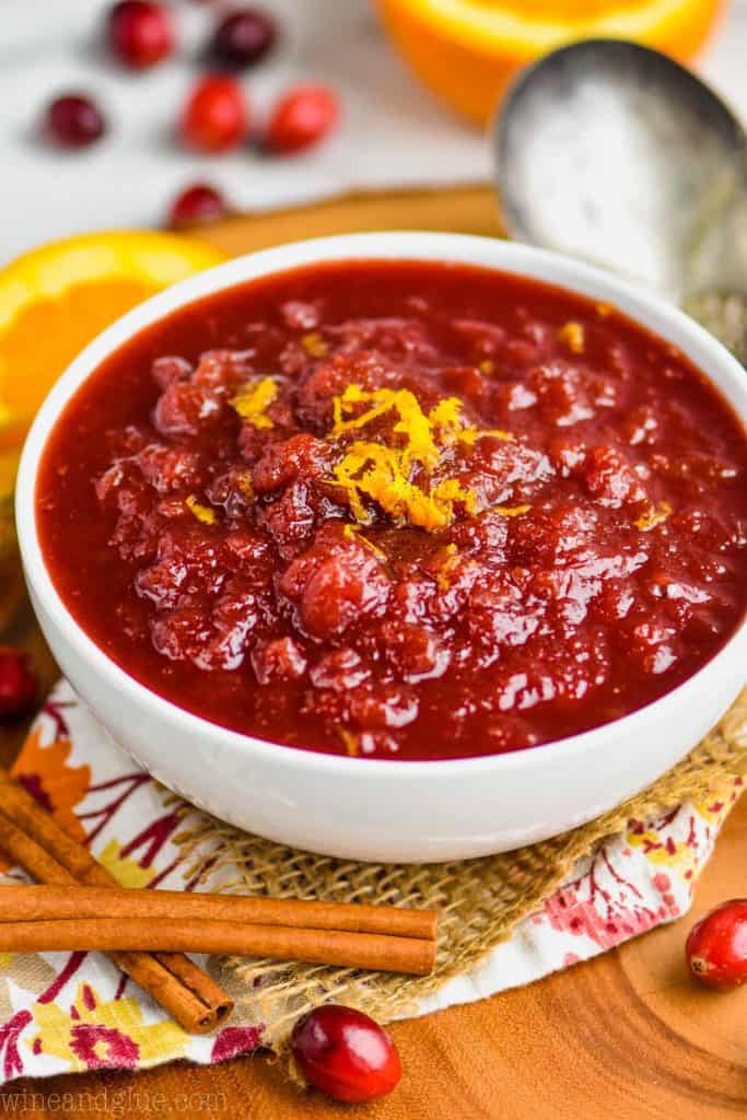 cranberry sauce in a white bowl with orange zest on top, sitting on a pieces of burlap and a fall napkin with cinnamon sticks and fresh cranberries around