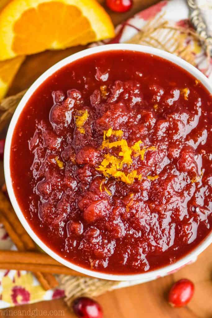 overhead of homemade cranberry sauce in a white bowl garnished with orange zest on a wood serving board with cinnamon sticks and fresh cranberries around