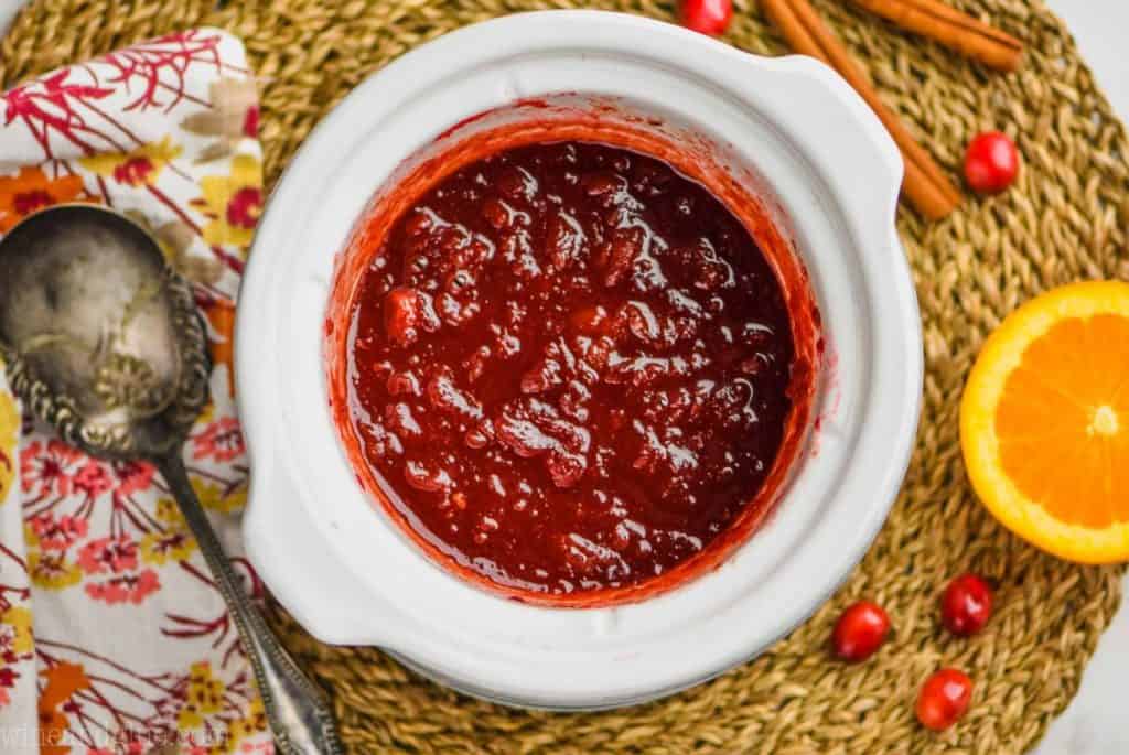 overhead of a small white slow cooker with fresh cranberry sauce in it, on a straw placemat with oranges, cinnamon sticks, and fresh cranberries around it