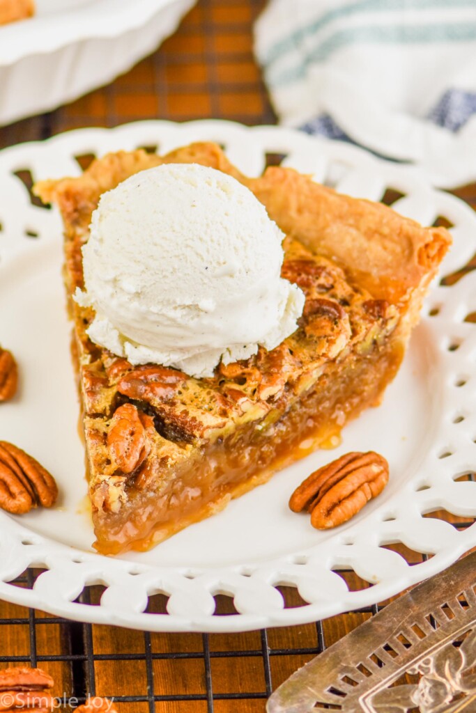 a piece of the best pecan pie on a fancy white plate with a large scoop of vanilla ice cream on top and the rest of the pie blurred in the background