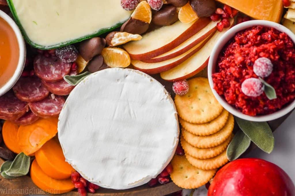 A close up photo of sliced persimmons, sliced apples, sliced meats, crackers, clementine slices dipped in chocolate, and candied nuts.  