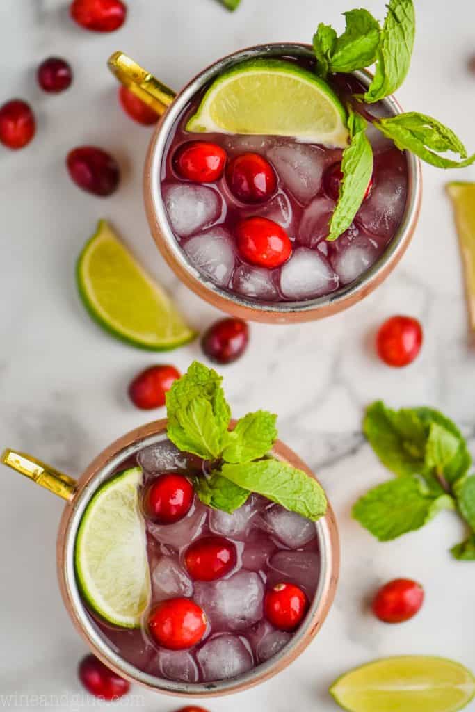overhead view of two copper mugs full of cranberry moscow mules, garnished with fresh cranberries, lime wedges, and sprigs of fresh mint