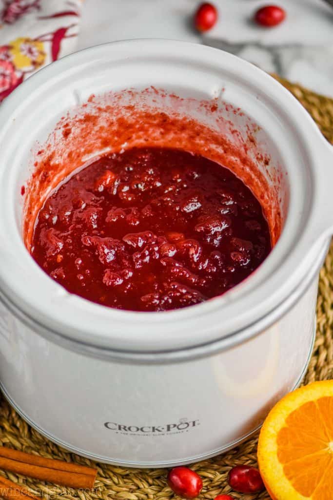 picture of crockpot cranberry sauce in a small white slow cooker with cinnamon sticks, fresh cranberries, and half an orange next to it.
