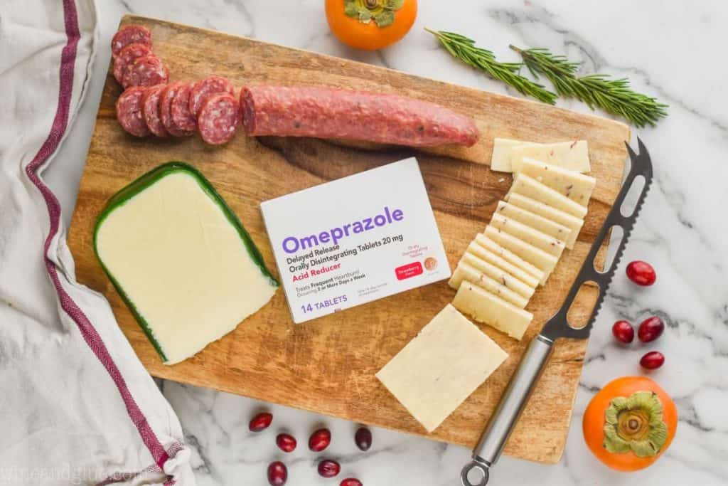An overhead photo of a square cheese board with  sliced meats and cheeses. In the middle of the cheese board, there is a box of Omeprazole (an acid reducer). 