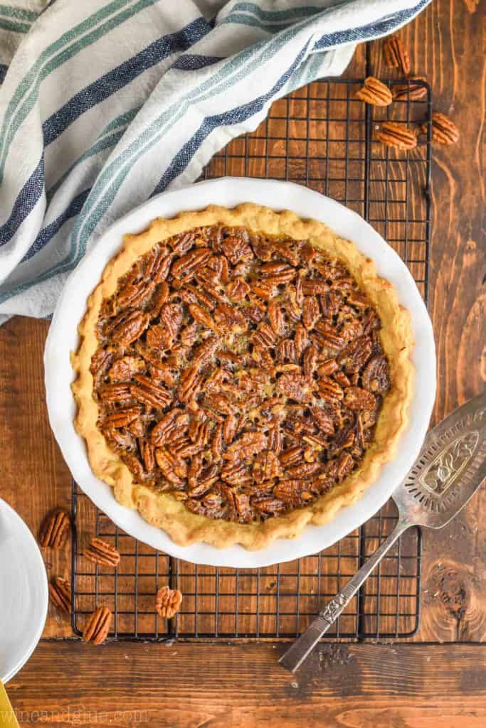 pulled back overhead view of the best pecan pie recipe in a white ceramic pie plate on a wire rack with an antique pie server next to it