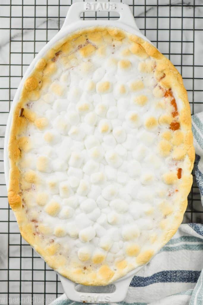 overhead view of a sweet potato casserole topped with marshmallows in a white oval casserole dish on a wire rack with a striped towel next to it