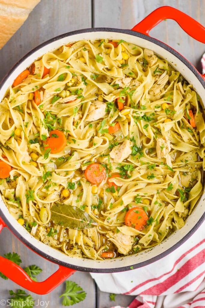 overhead view of a red handled stock pot full of turkey noodle soup - long noodles, pieces of turkey, a bay leaf, carrots, and corn are visible 