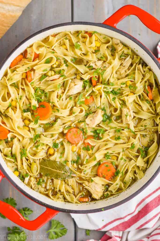 overhead view of a red handled stock pot full of turkey noodle soup - long noodles, pieces of turkey, a bay leaf, carrots, and corn are visible 