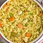 overhead view of a red handled stock pot full of turkey noodle soup - long noodles, pieces of turkey, a bay leaf, carrots, and corn are visible