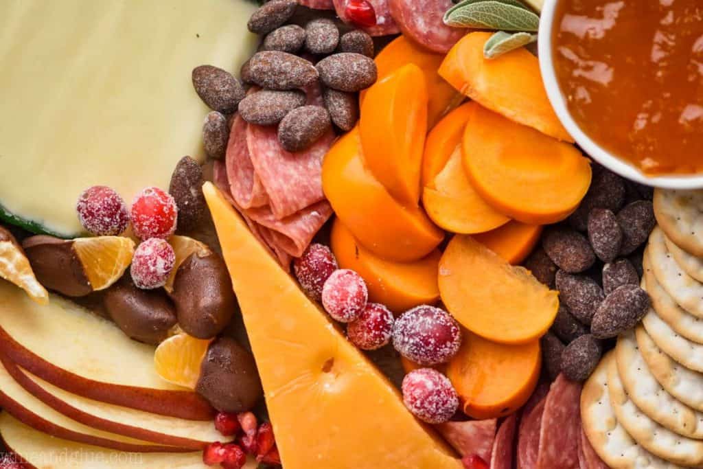 A close up photo of sliced persimmons, sliced apples, sliced meats, crackers, clementine slices dipped in chocolate, and candied nuts.  