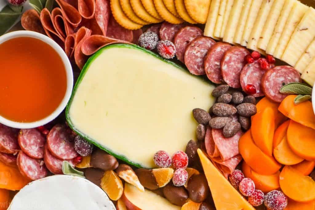 A close up photo of sliced persimmons, sliced apples, sliced meats, crackers, clementine slices dipped in chocolate, and candied nuts.  