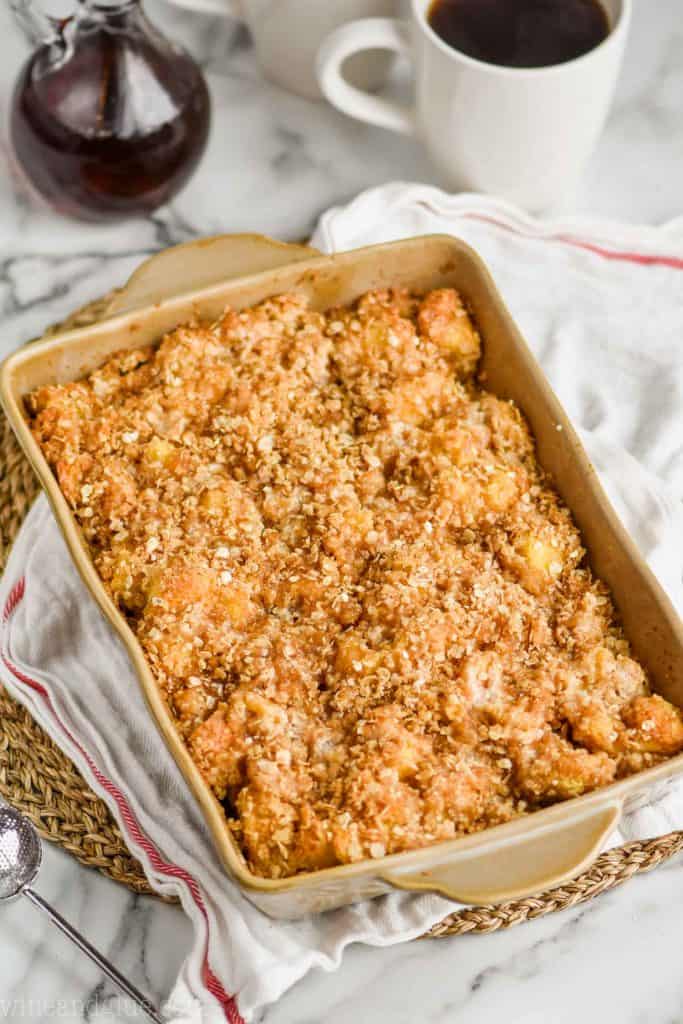 overhead view of baked French toast casserole in a brown ceramic baking dish on a cloth napkin and straw circle placemat with bottle of syrup