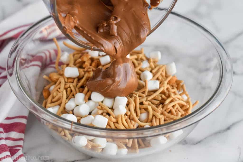 The mixture of melted butterscotch and chocolate is being poured over the peanuts, marshmallows, and the chow mien noodles mixture. 