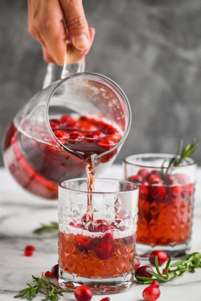 hand holding a pitcher of sangria pouring it into a glass with a full glass garnished with fresh cranberries in the background