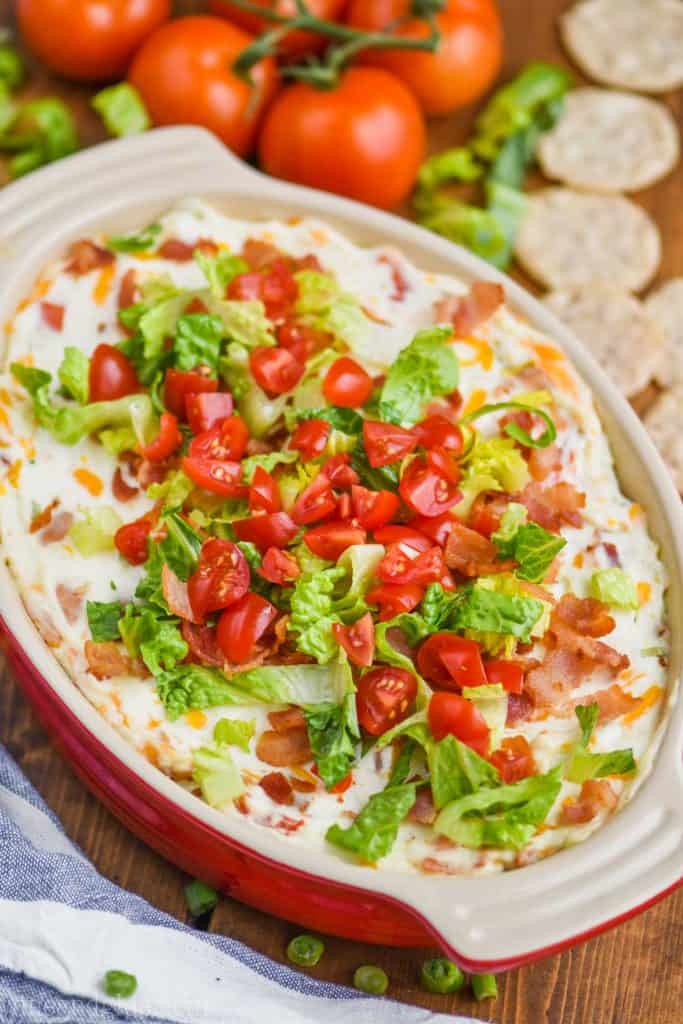 pulled back view of a red ceramic baking dish filled with blt dip recipe, topped with chopped tomatoes and lettuce, with whole tomatoes in the background