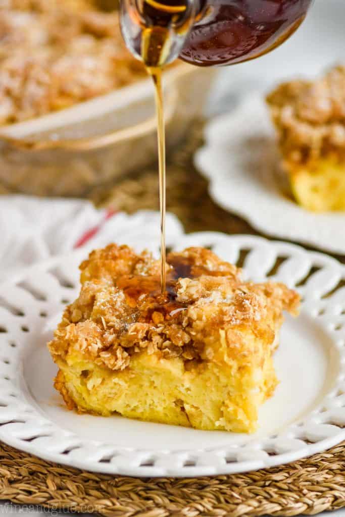 a square piece of French toast casserole on a fancy whit plate with streusel on top, and syrup being poured on