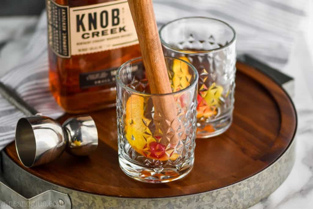 a small tumbler glass with fruit in the bottom being muddles, with a whiskey bottle in the background