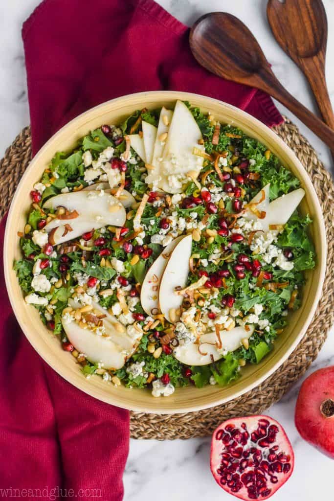 overhead view of a wooden serving bowl full of winter salad recipe with sliced pears, kale, blue cheese, crispy leeks, and pine nuts on a maroon cloth napkin with a pomegranate cut in half and a whole pomegranate next to it