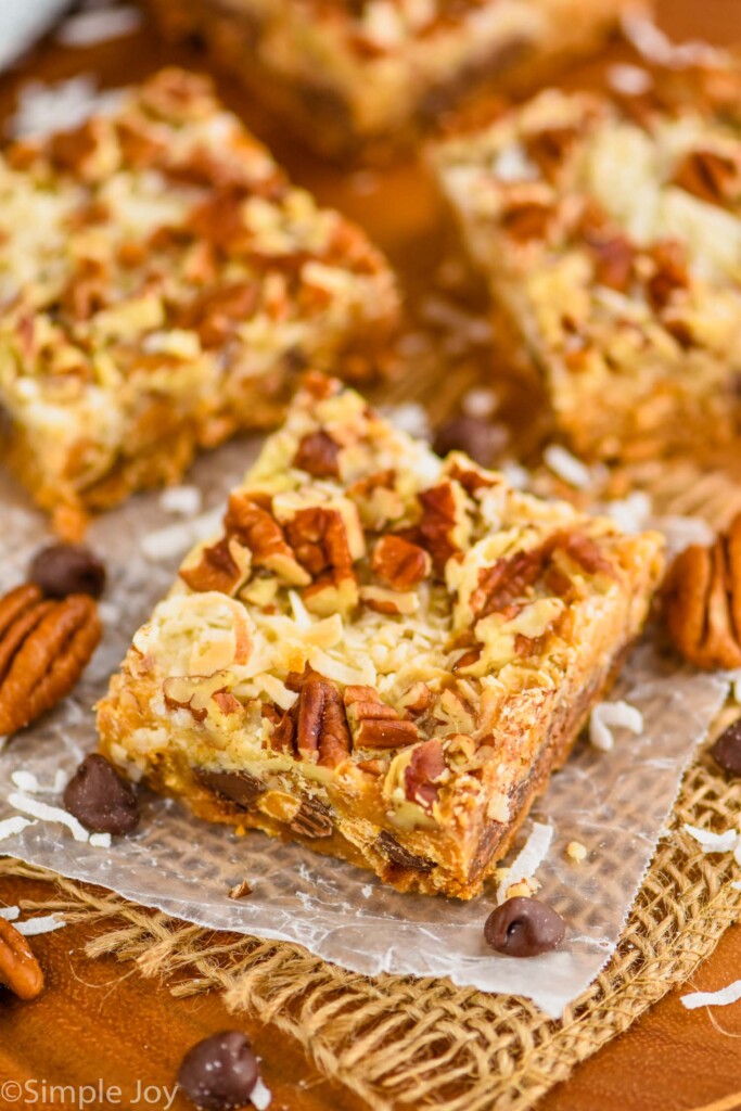 four seven layer bars sitting on parchment paper on a wood board with coconut, pecans, and chocolate chips around them