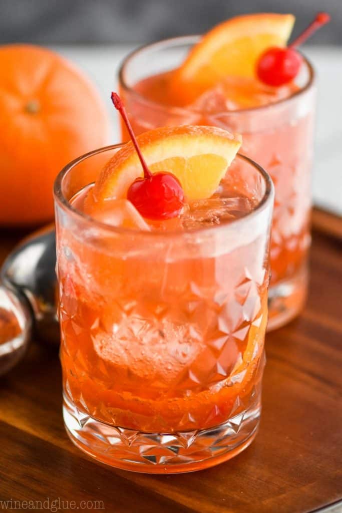 an old fashioned cocktail on a wood tray, garnished with an orange wedge and a cherry, with another right behind it