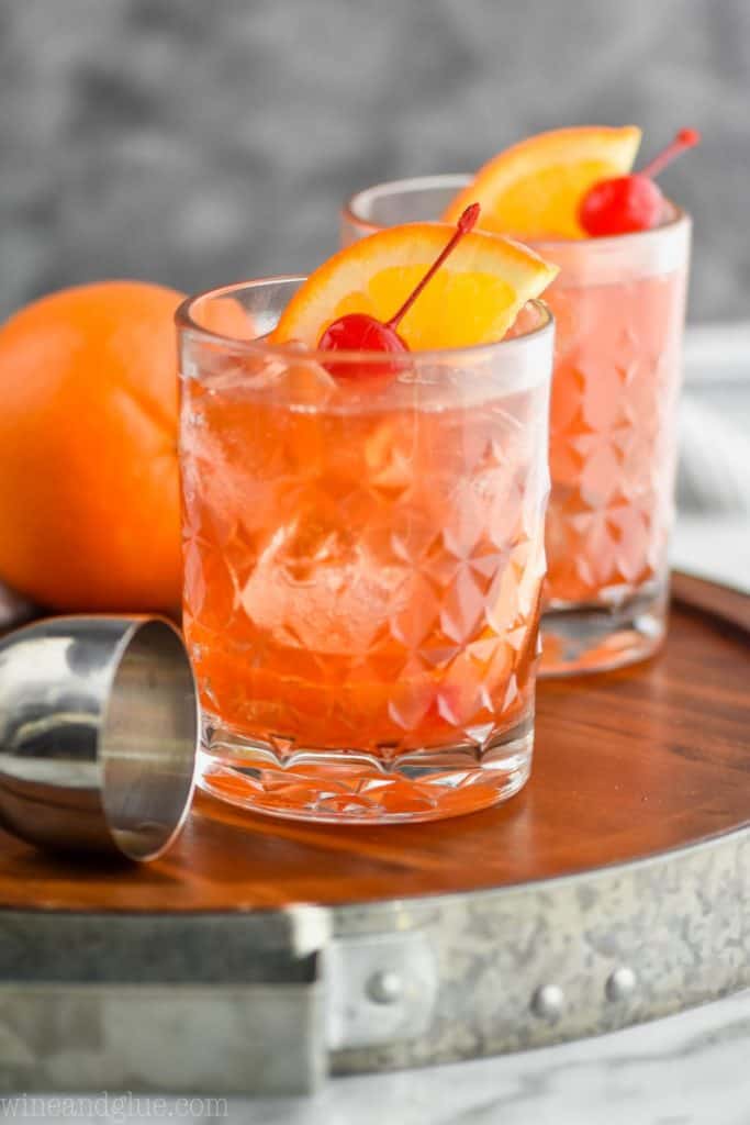 two small glass tumblers containing old fashioned drink recipe on a steel rimmed wood tray containing an old fashioned drink