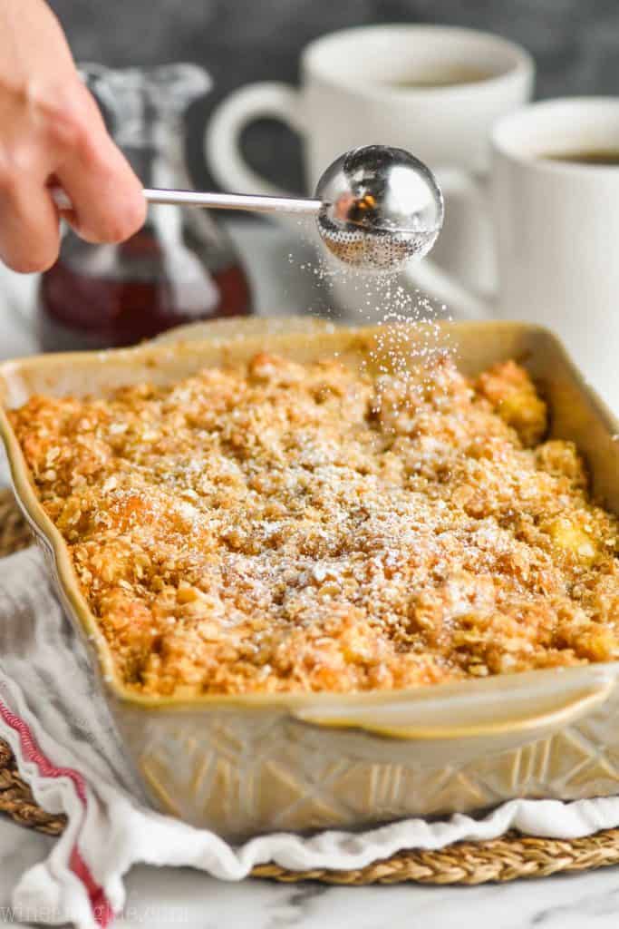 silver sphere powdered sugar wand, dusting powdered sugar over overnight French toast casserole in a ceramic baking dish on a cloth napkin