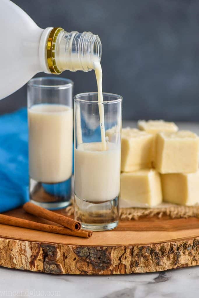 two tall shot glasses on a wood board with fudge next to them and rum Chata being poured into one