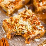 up close overhead picture of a seven layer bar on a piece of parchment paper with a bite missing, coconut shreds and pecan pieces visible on the top of the gooey bar